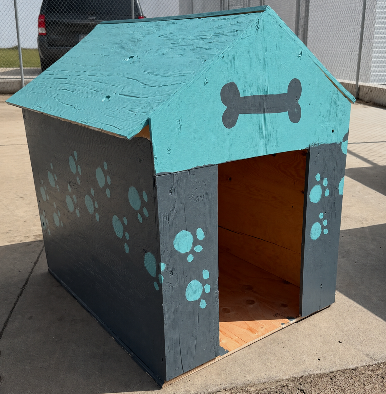 A hand-painted teal and grey Project Archie doghouse with paw prints and a bone motif, donated to a local dog shelter