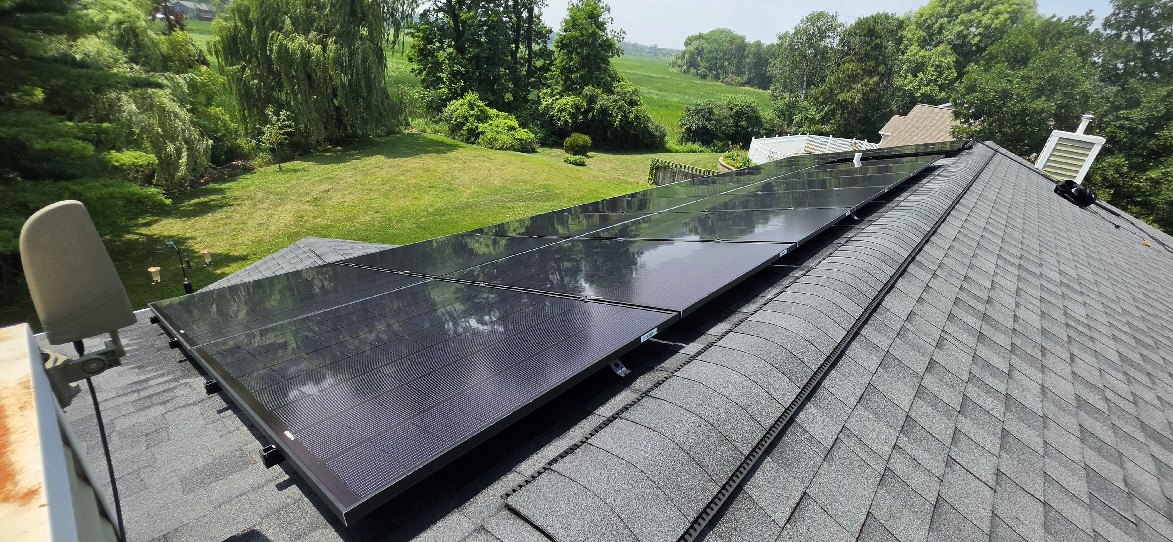Same rural Southern Wisconsin residential solar array photographed from the opposite roof end after cleaning, with tree canopies reflected in the glass and open cornfields on the horizon