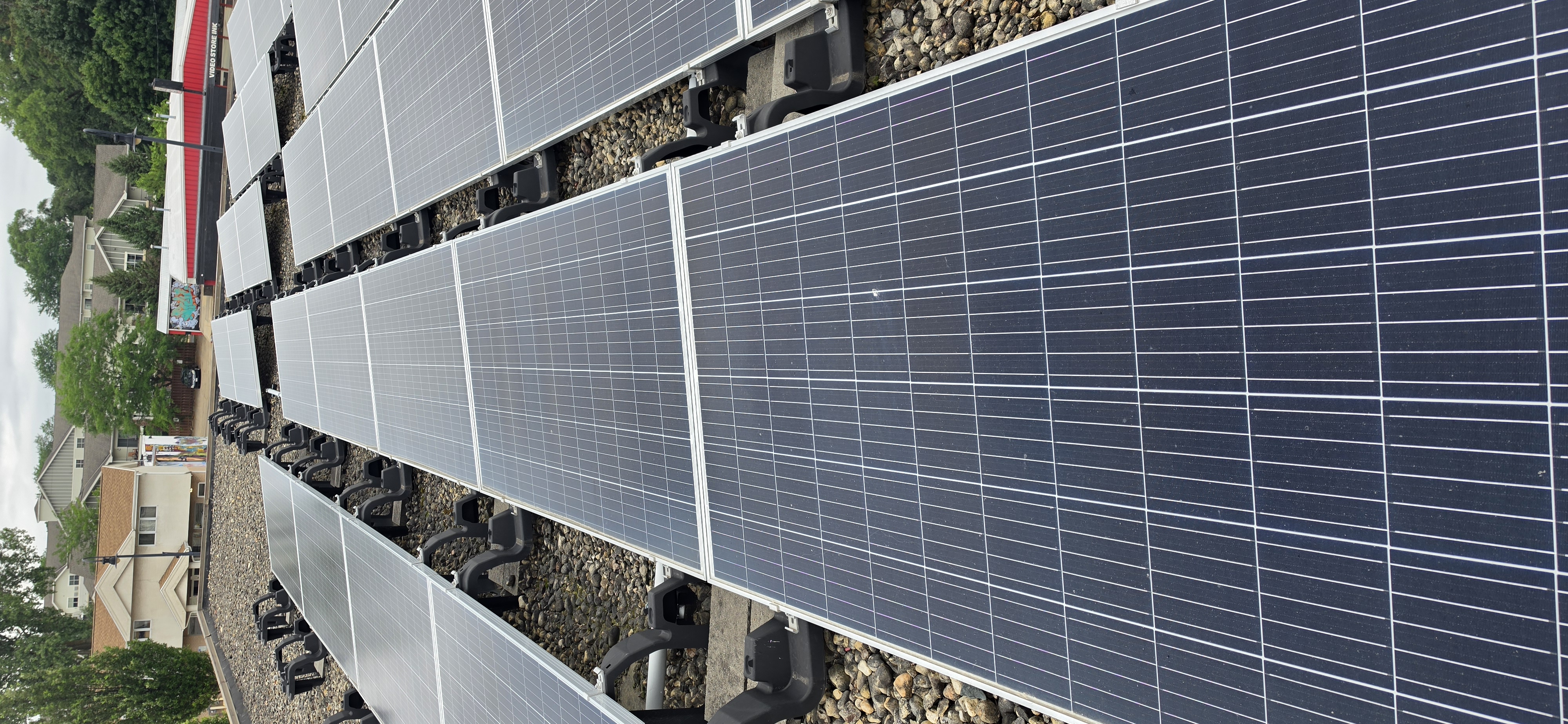 Wide-angle after photo of the freshly cleaned commercial flat-roof solar array — rows of ballasted panels running toward the residential neighborhood beyond, each panel now showing deep uniform blue with visible reflection detail