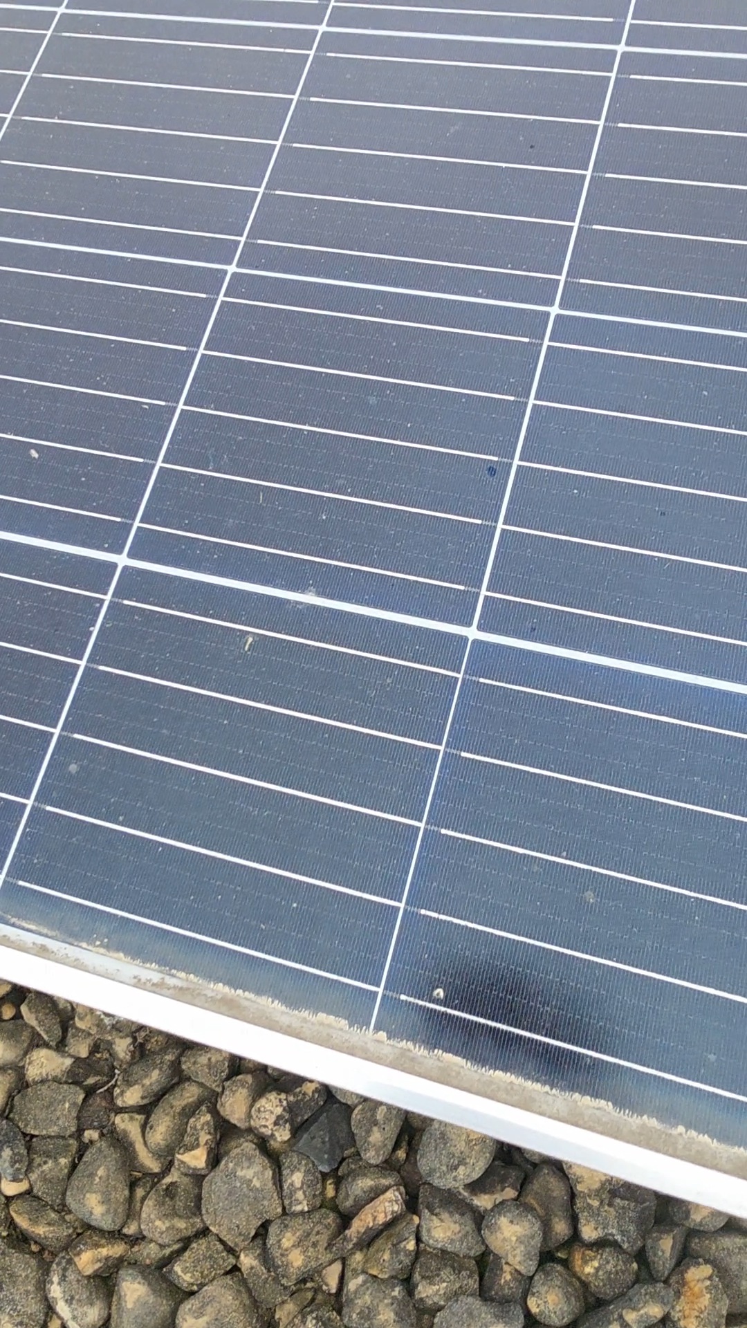 Extreme close-up of the same commercial solar panel showing a dust-and-particulate film along the bottom edge before cleaning, with ballast gravel and rocks visible around the panel frame