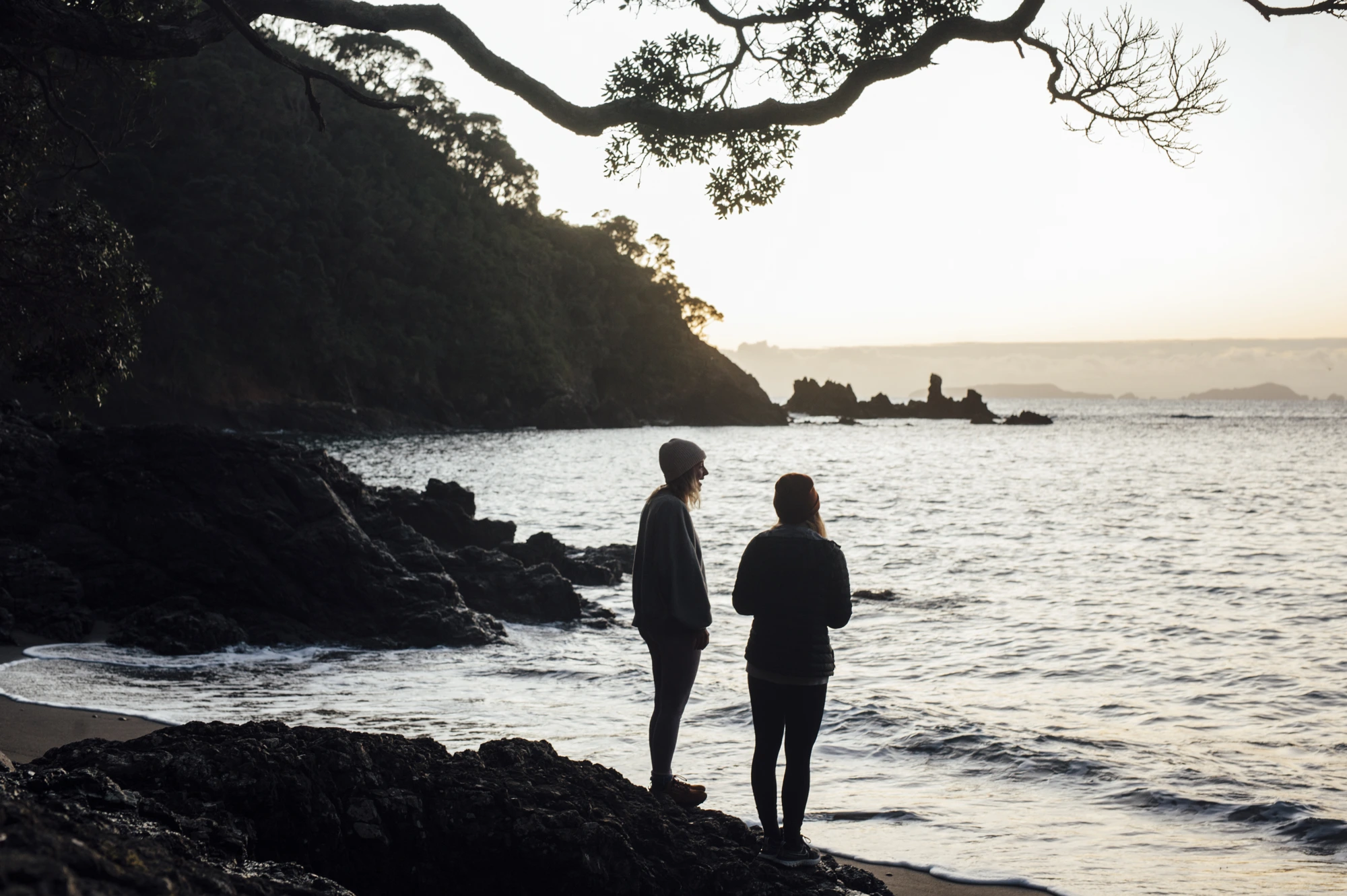 Two women standing together on the rocks at Whananaki, looking out to sea at dusk
